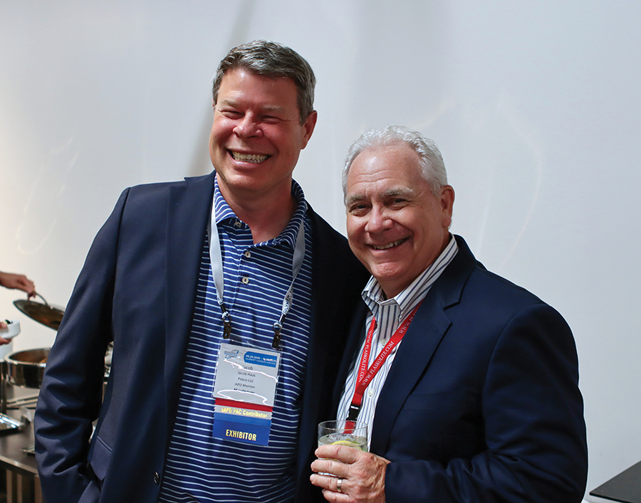 Two men wearing conference lanyards and suits smiling together while holding a drink.